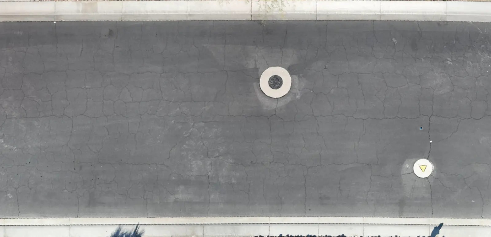 Top-down drone photo of an asphalt road between two curbs, with a manhole cover and a round marker in the lane and cracking that is hard to see by eye.