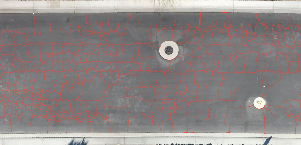 Asphalt street between two curbs from directly above, with every crack traced in red from curb to curb, including the cracking around a manhole cover