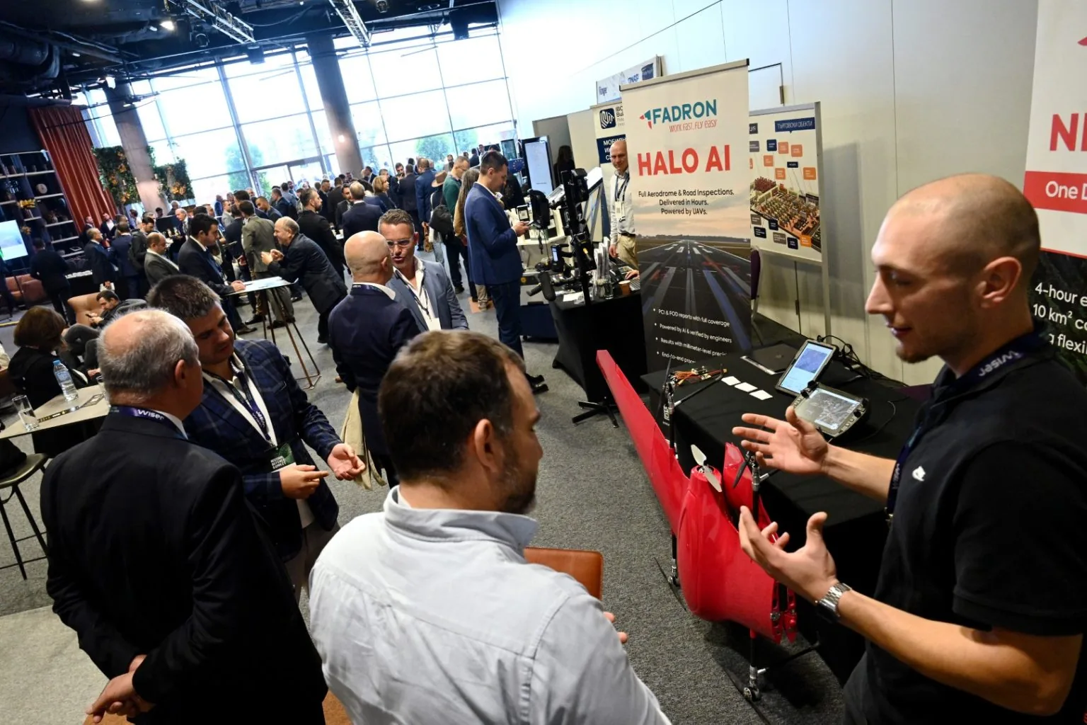 Visitors talking with a Fadron engineer beside a red Nimbus on the Fadron stand at a crowded industry exhibition