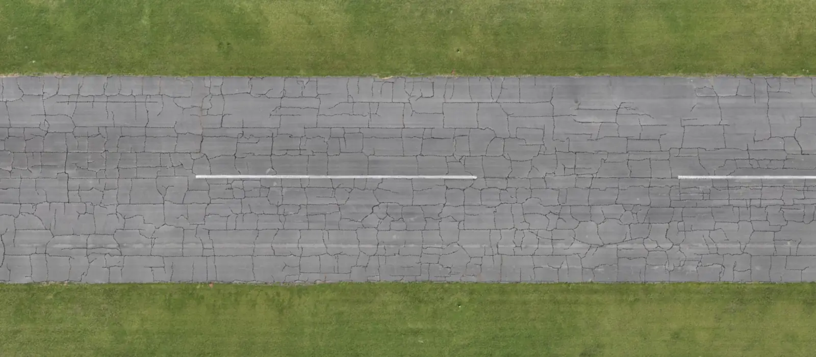 Ortomosaico de dron de una pista de asfalto visto desde una perspectiva cenital, con la pintura de la línea central atravesando una densa red de grietas en bloque y césped a ambos lados.